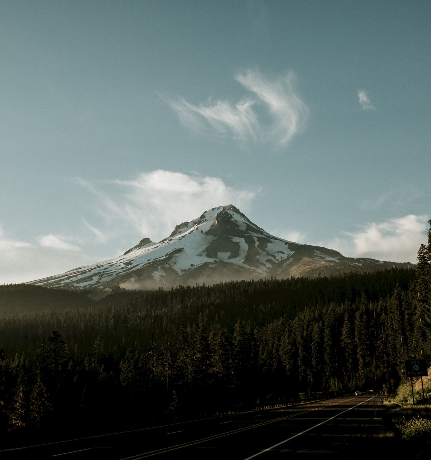 A snow-capped mountain is surrounded by a dense forest. A winding road cuts through the forest, heading towards the mountain.