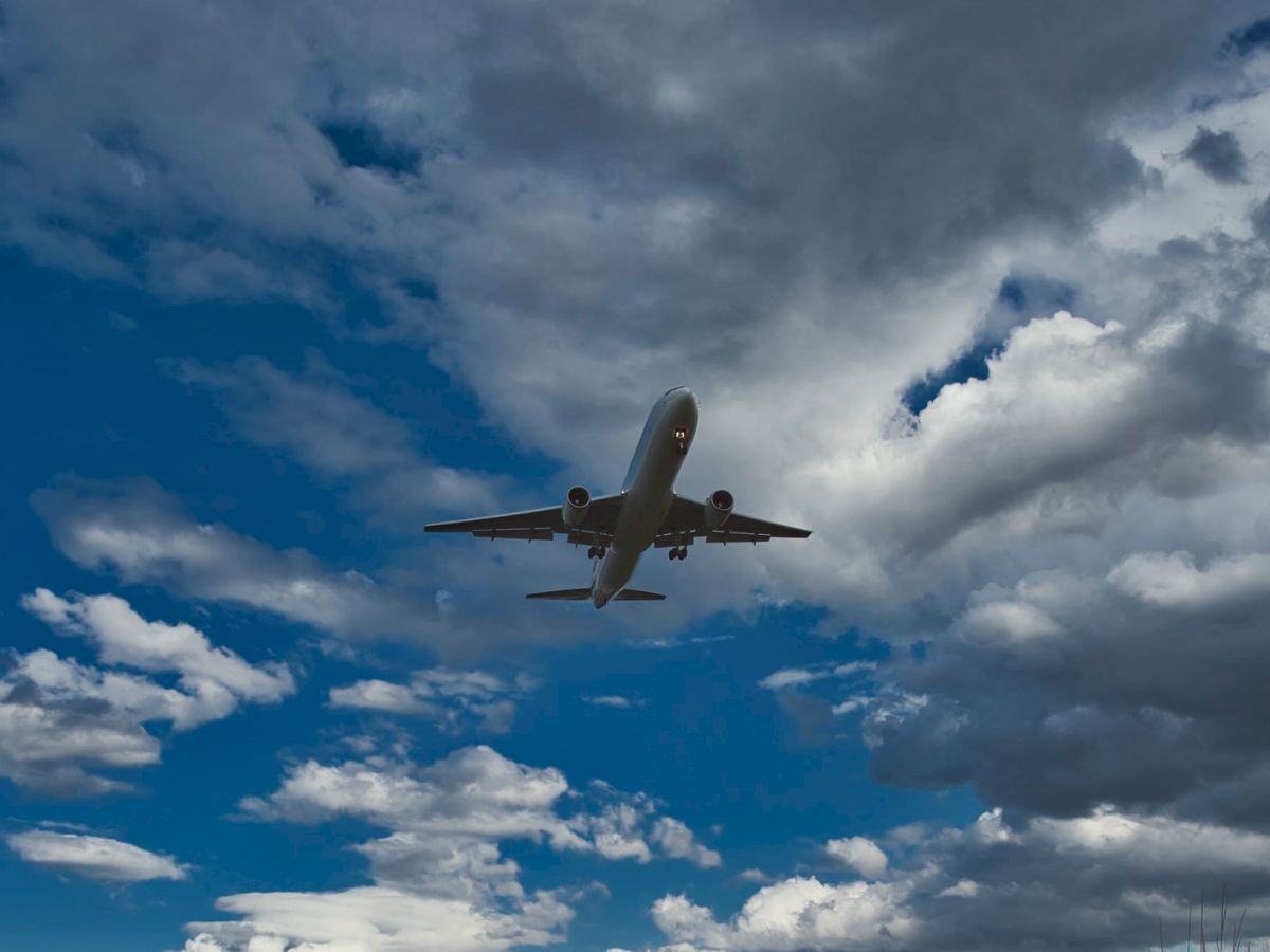 A plane flying low over a path by a body of water, with a grassy hill on the right side under a partly cloudy sky, ending the sentence.