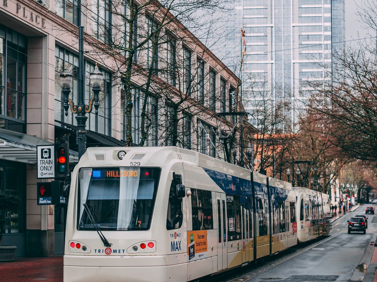 A modern light rail train travels down a wet city street with tall buildings in the background and a few pedestrians on the sidewalks.