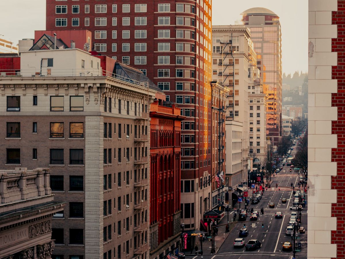 The image shows a city street lined with tall buildings and traffic. The sun appears to be setting or rising, casting a warm glow over the scene.