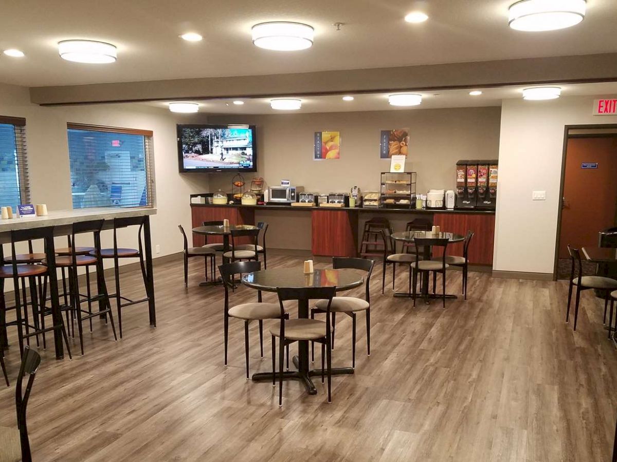 The image shows a modern, well-lit dining area with various tables and chairs, a counter with breakfast items, and a wall-mounted TV displaying a sports game.