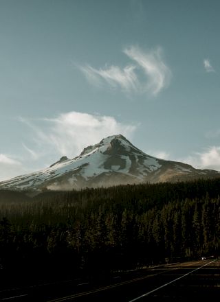 A snow-capped mountain peak under a blue sky, surrounded by dense forests and flanked by a winding road on the right side of the image.