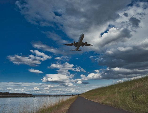 An airplane is flying in the sky over a pathway next to a body of water; the sky is partly cloudy with patches of blue showing.