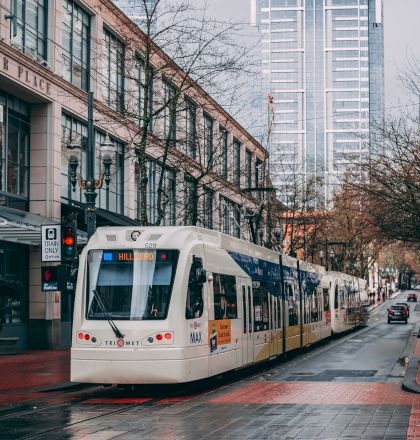 A white tram runs along a wet urban street lined with modern buildings and trees, leading toward a tall skyscraper in the background.