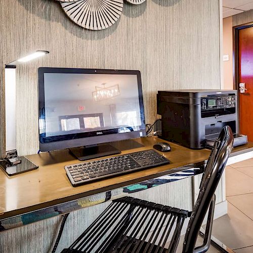 The image shows a small office setup with a computer, keyboard, monitor, desk lamp, and printer on a desk, next to a chair in a room.