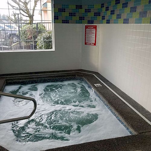 An indoor hot tub with bubbly water is situated in a tiled room with a window and a railing for entry. A sign is visible on the wall.