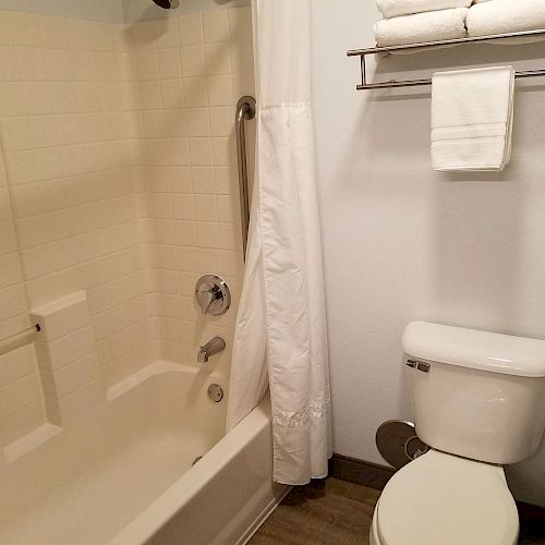 A bathroom with a shower-tub combo, white curtain, toilet, and a towel rack holding towels on the wall above the toilet, ending the sentence.