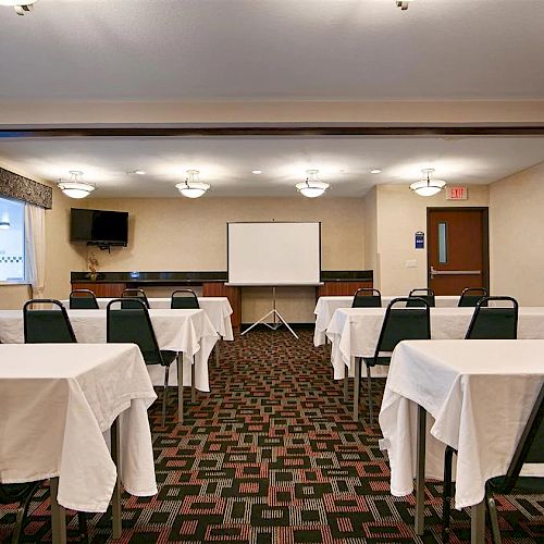 A small conference or meeting room with rows of tables and chairs, a projector screen, and a television mounted on the wall at the front of the room.