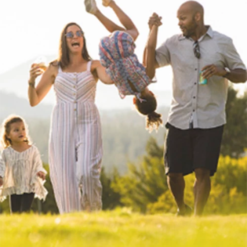 A family of four enjoys a sunny day outdoors. The father is swinging one child upside-down while the mother and another child smile and walk alongside.