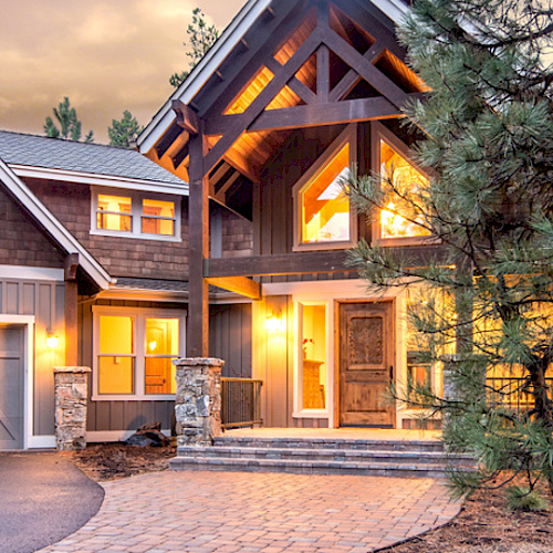 A beautiful, illuminated large house with stone accents, a spacious driveway, and a garage, surrounded by trees, under a twilight sky.