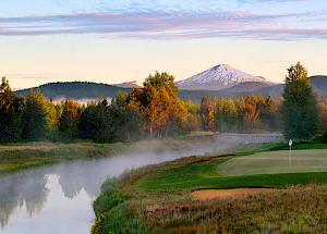 A scenic view of a golf course near a river with mist, surrounded by trees and mountains with a snow-capped peak under a colorful sky.