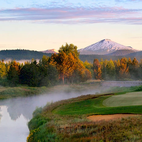 A scenic view of a golf course near a river with mist, surrounded by trees and mountains with a snow-capped peak under a colorful sky.