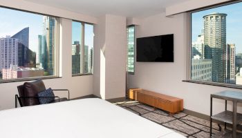 A modern bedroom with large windows showing a cityscape, a wall-mounted TV, a chair, a low table, and minimalistic decor.
