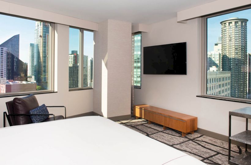 A modern bedroom with large windows showing a cityscape, a wall-mounted TV, a chair, a low table, and minimalistic decor.