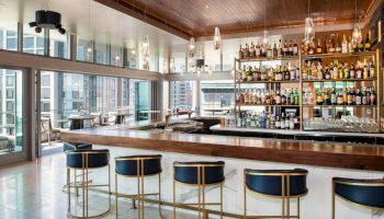 The image shows a modern bar with blue stools, a wooden countertop, and shelves stocked with various liquor bottles, set in a room with large windows.
