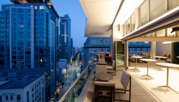 A modern cityscape shows tall buildings at dusk, a furnished balcony overlooking the street with tables, chairs, and open doors leading inside.
