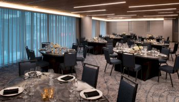 A modern, elegantly set dining room with round tables, black chairs, table settings, flowers, and candles, illuminated by overhead lights and natural light.