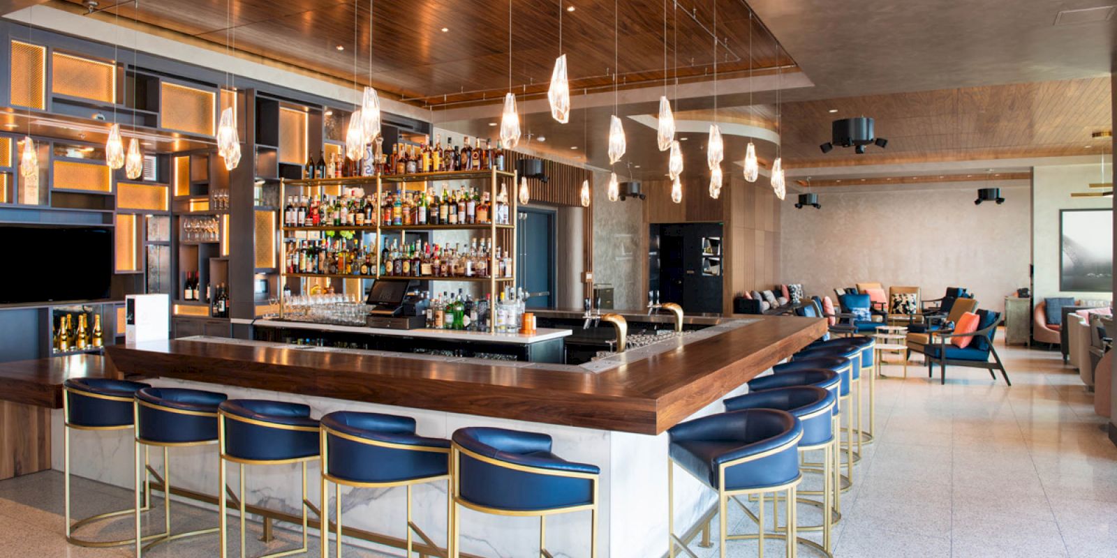 A stylish modern bar with blue cushioned barstools, hanging light fixtures, and a well-stocked liquor shelf behind the counter.