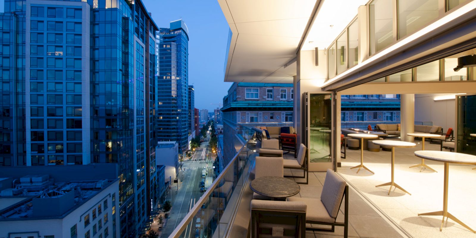 A modern balcony with tables and chairs overlooks a city street lined with tall buildings. The cityscape is illuminated in the evening.