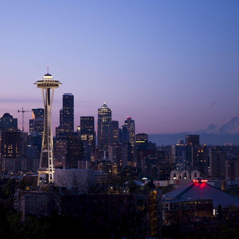 A twilight view of Seattle's skyline, featuring the Space Needle, numerous skyscrapers, and Mount Rainier in the background against a dusky sky.