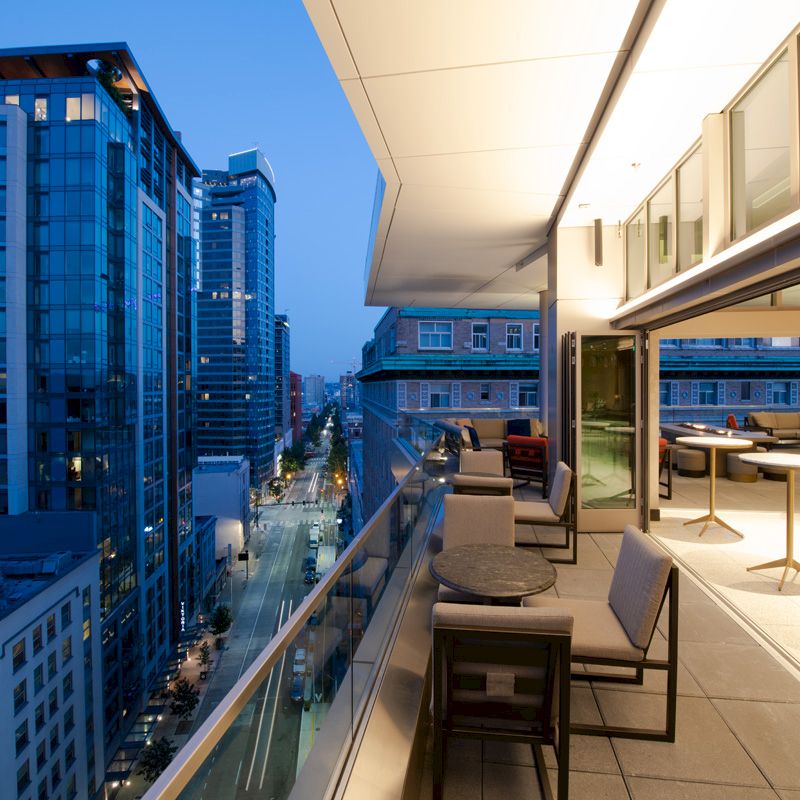 A modern cityscape view from a high-rise balcony at dusk, featuring outdoor furniture and tables, with buildings lining a street below.
