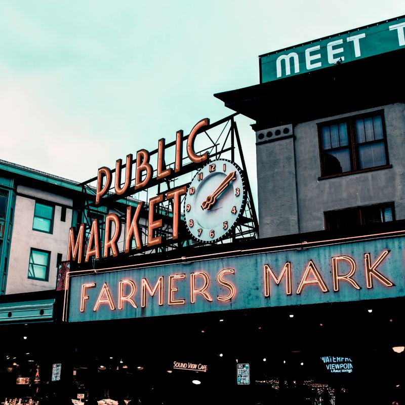 The image shows a neon sign for a "Public Market" with a clock, and another sign below saying "Farmers Market" in front of buildings.