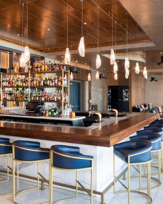 A modern bar with blue stools, a wooden counter, hanging lights, and a well-stocked liquor shelf, set in a stylish lounge area.