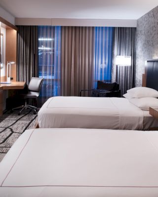 A modern hotel room featuring two beds, a wall-mounted TV, a study desk with a chair, and large windows with curtains and ambient lighting around.