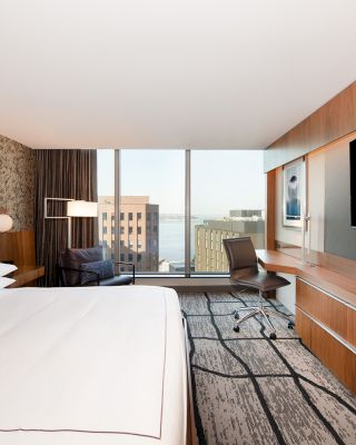 A modern hotel room with a large bed, wall-mounted TV, desk, chair, and a window offering a city view with water in the background.