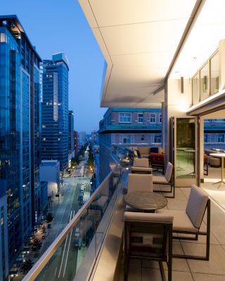 A modern cityscape at dusk with tall buildings and an elevated outdoor terrace featuring seating and tables overlooking the street below.