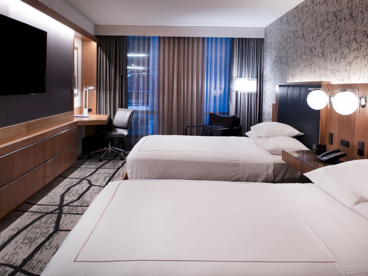 A hotel room with two beds, nightstands, a TV, a desk, a chair, and modern decor featuring neutral tones and patterned wallpaper.