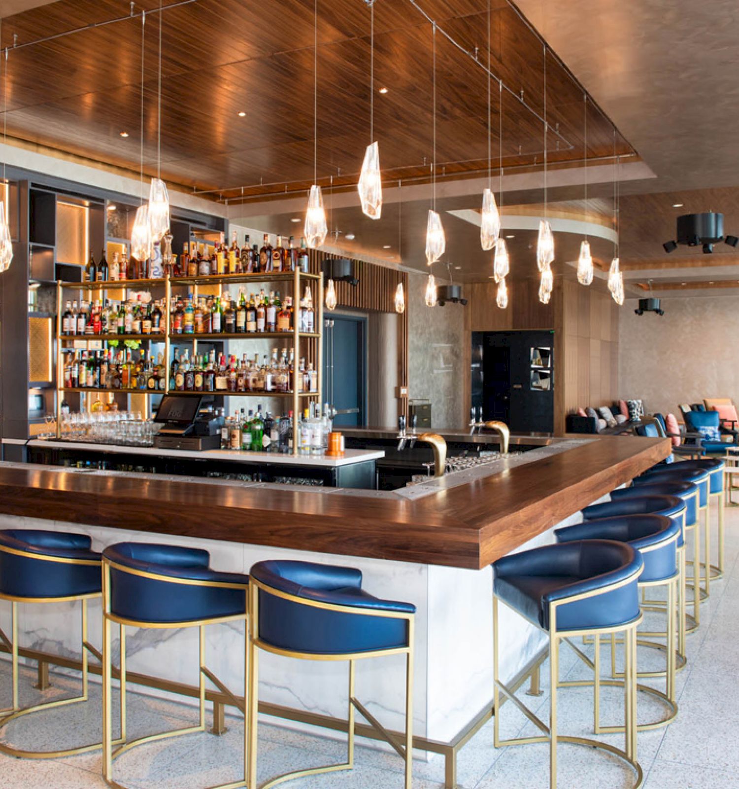 The image shows a modern bar with blue bar stools, wooden countertops, hanging lights, and a well-stocked shelf of bottles. It has a chic interior.
