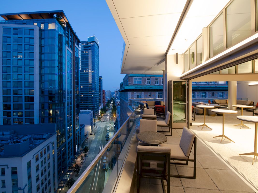 A modern city view from a balcony at dusk, featuring outdoor furniture, tall buildings with glass exteriors, and lighted street below.