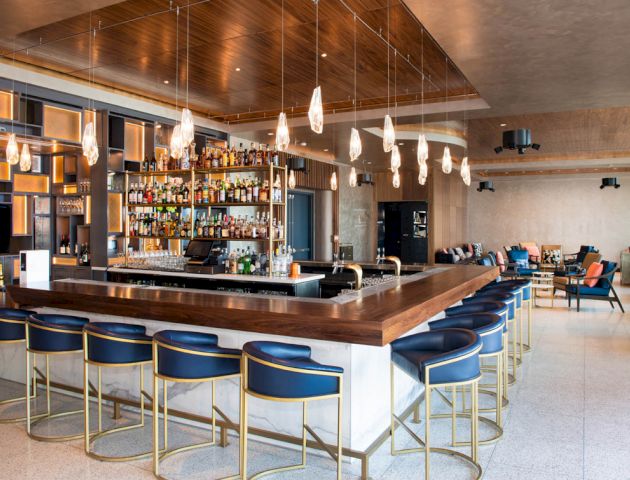 This image shows a modern bar with a well-stocked liquor shelf, bar stools, pendant lighting, and a lounge area in the background.