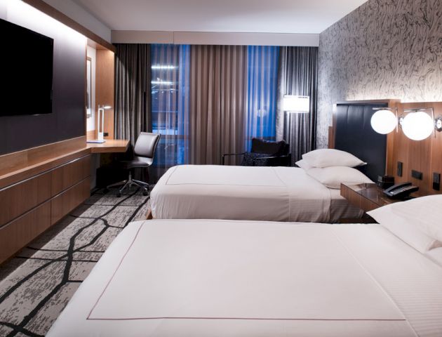 The image shows a modern hotel room with two neatly made beds, a wall-mounted TV, a desk by the window, and contemporary decor.