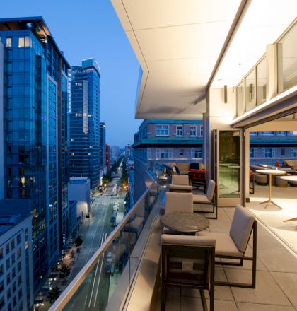 A modern balcony at dusk overlooks a city street lined with tall buildings, featuring comfortable seating and round tables under warm lighting.