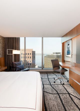 The image shows a modern hotel room with a large bed, a big TV, a desk with a chair, and a window view of city buildings and water.
