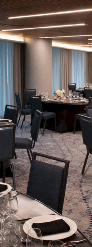 The image shows a modern banquet hall with round tables, set with silverware, glassware, and black napkins, ready for an event or dining.
