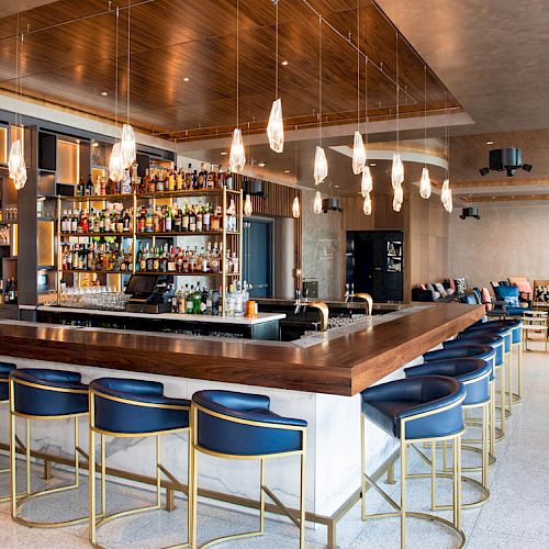 The image shows a modern bar area with a wooden countertop, blue chairs, hanging lights, and shelves stocked with various bottles of alcohol.