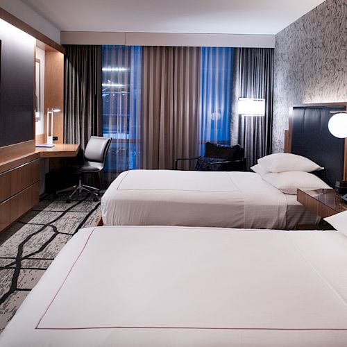 A modern hotel room with two neatly made beds, a desk with a chair, a large TV on the wall, and contemporary decor with patterned wallpaper.
