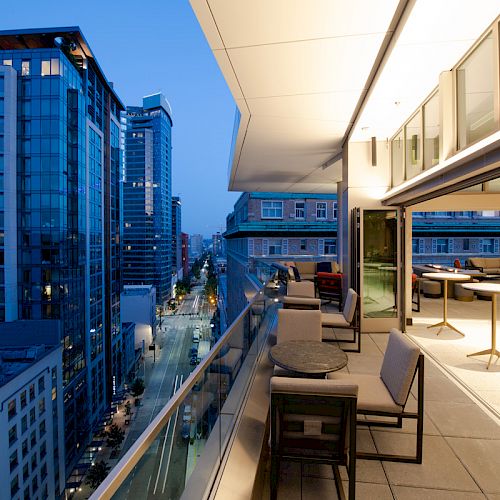A modern balcony with tables and chairs overlooks a city street lined with tall buildings during dusk.