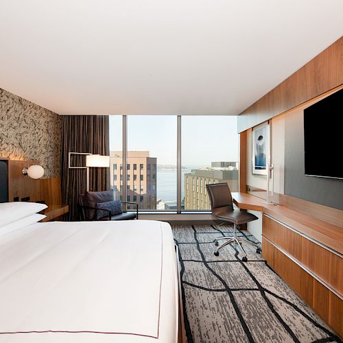 The image shows a modern hotel room with a large bed, a desk, and a flat-screen TV. The window offers a view of nearby buildings and the water.