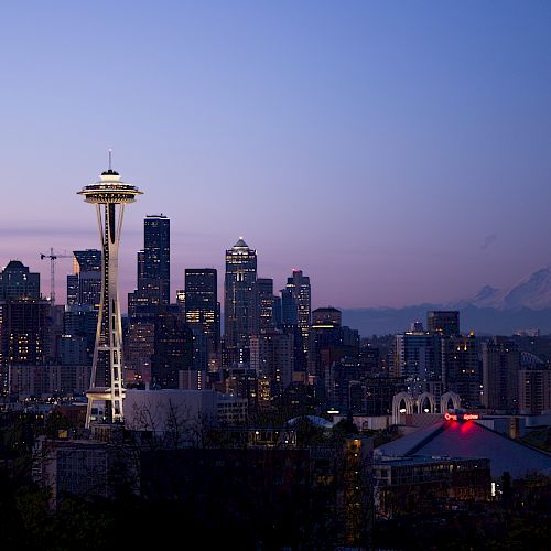 City skyline at dusk featuring a prominent tower and various buildings, with a mountain in the background and a colorful evening sky.