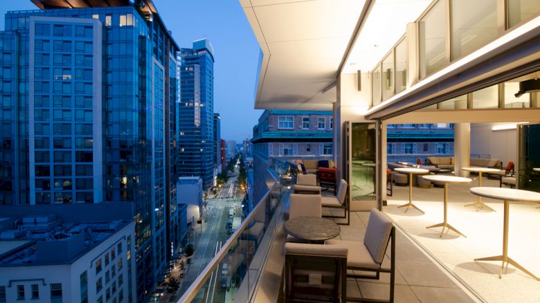 This image features a modern cityscape at dusk, viewed from a balcony with sleek outdoor furniture, overlooking tall buildings and a lit-up street.