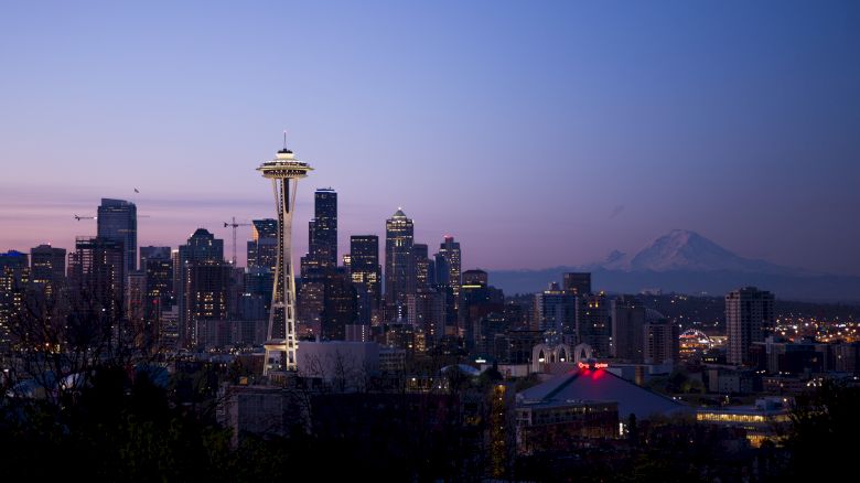 A cityscape at dusk featuring the Seattle skyline, with the Space Needle prominently visible and Mount Rainier in the background.