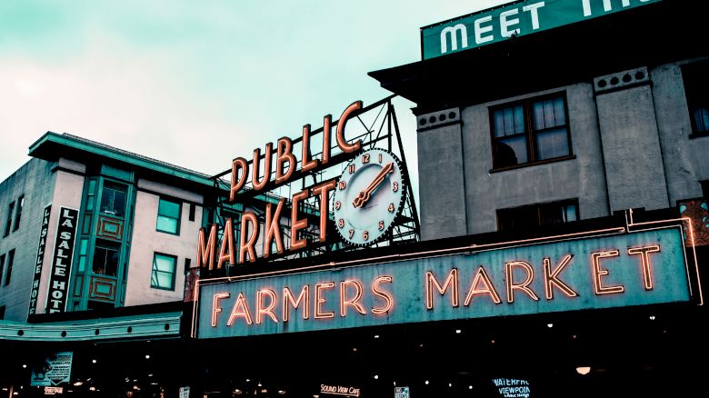 The image shows a public market with neon signs reading "Public Market" and "Farmers Market" on buildings, next to a large clock.
