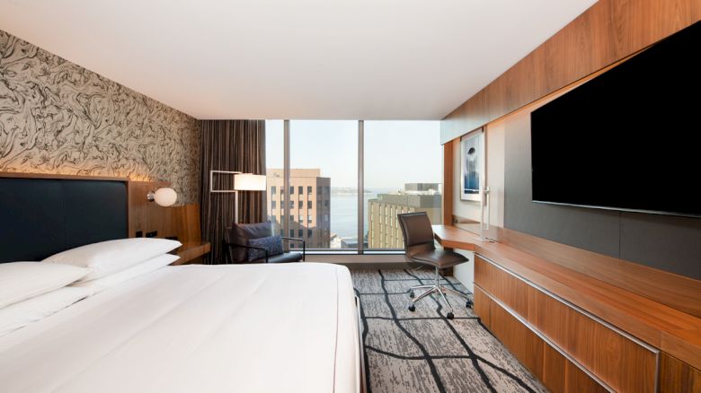 A modern hotel room with a large bed, flat-screen TV, desk, and floor-to-ceiling windows offering a city view.