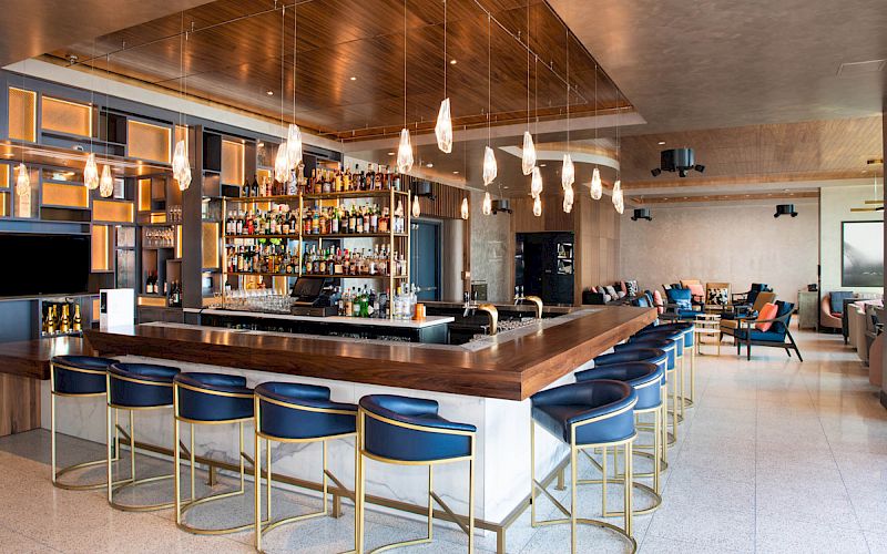 This image shows a modern bar with sleek wooden countertops, blue bar stools, hanging lights, and a well-stocked shelf of liquor bottles.