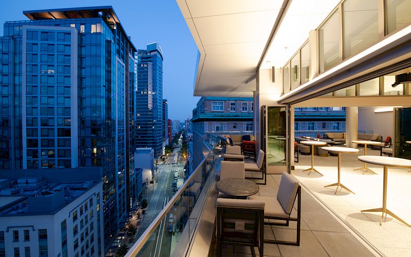 A modern cityscape with tall buildings is visible from a balcony with outdoor seating and tables at dusk.