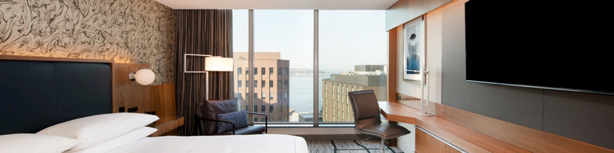 A modern hotel room featuring a large bed, a desk, a chair, a TV, and a window with a city view in the background.